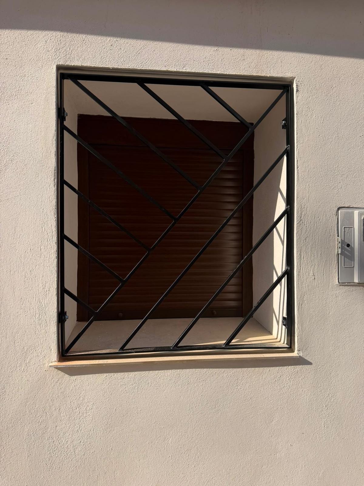 reja diagonal negra instalada en ventana con persiana marrón, fachada blanca al sol