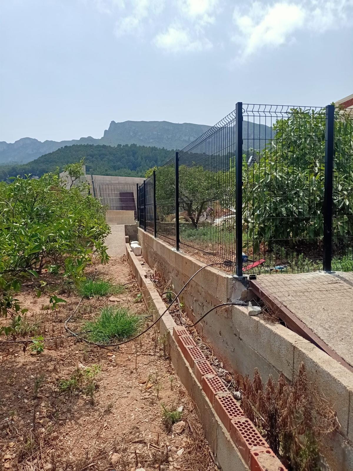 valla de malla electrosoldada negra sobre muro de bloques, con montaña al fondo
