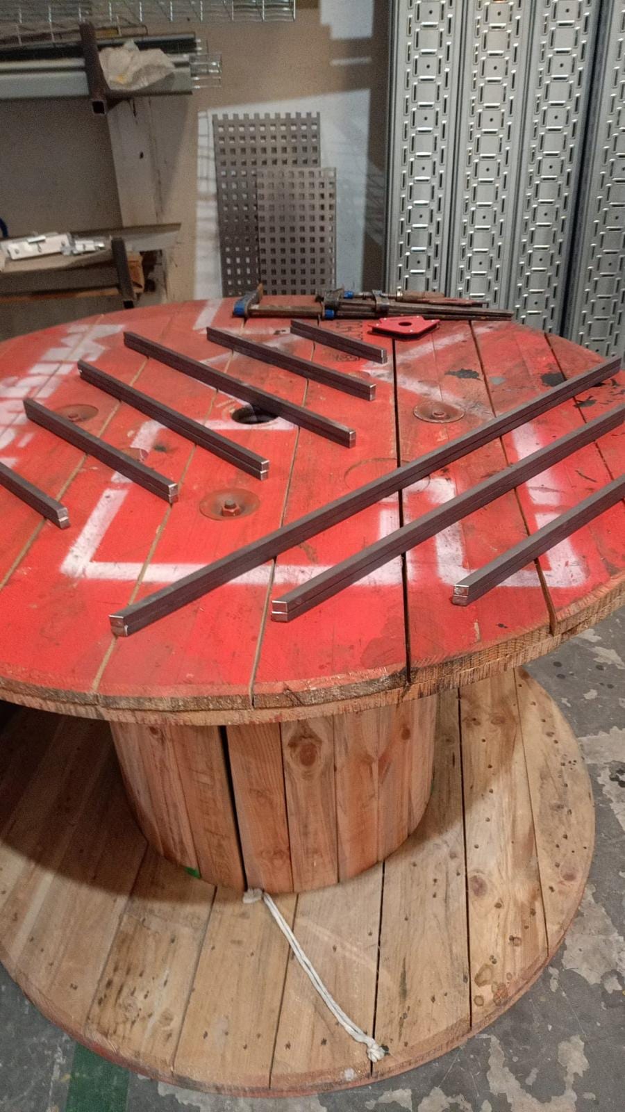 Barras de tubo cuadrado ya cortadas a inglete y colocadas en diagonal sobre una bobina de cable que hace de mesa en el taller, con sargentos al fondo.