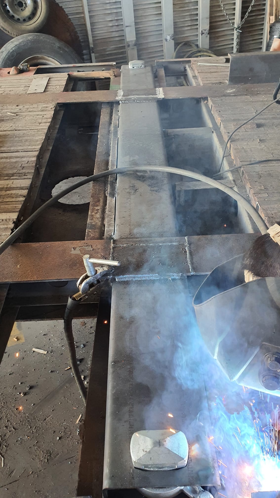 Soldador con pantalla uniendo travesaños al chasis de un remolque con anclajes de contenedor, entre chispas y humo en la nave.